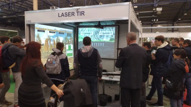 Перед двумя большими экранами люди стоят в очереди к бизнес аттракциону лазерный тир